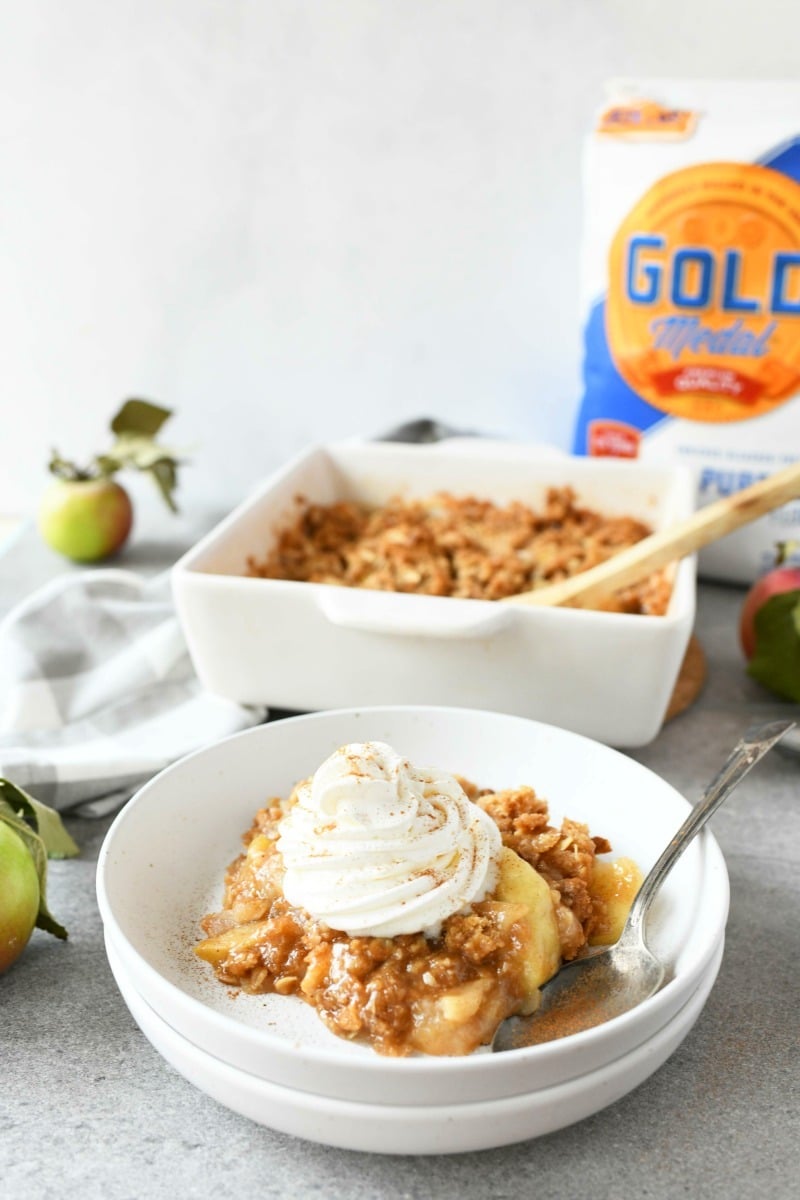 Gold Medal Apple Crisp with whipped topping and a spoon. A Flour bag in in the background on a gray table.