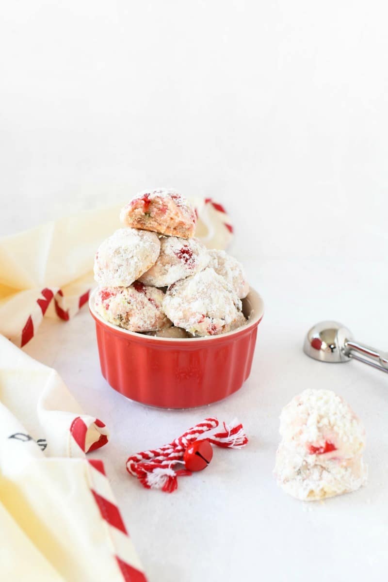 Pistachio Cherry Butter Cookies on a white table with a holiday napkin, cookie scoop, and bow.