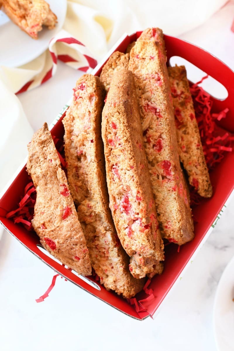 Toasted Cherry Almond Biscotti in a red baking box with red paper shreds on a white table. 