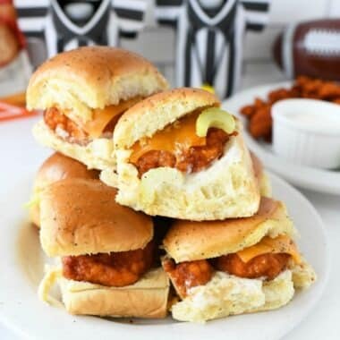 Buffalo Chicken Sliders are stacked on a white plate. There are black and white cozies in the background.