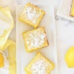 Lemon Bars cut on a white rectangle dish with a yellow napkin and fresh lemon nearby.