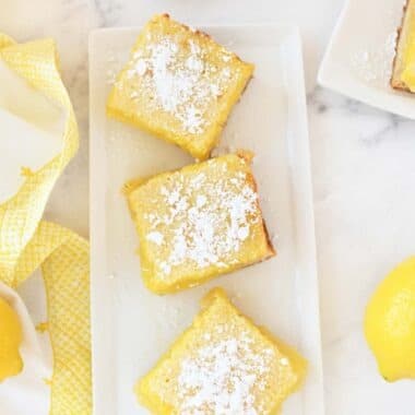 Lemon Bars cut on a white rectangle dish with a yellow napkin and fresh lemon nearby.