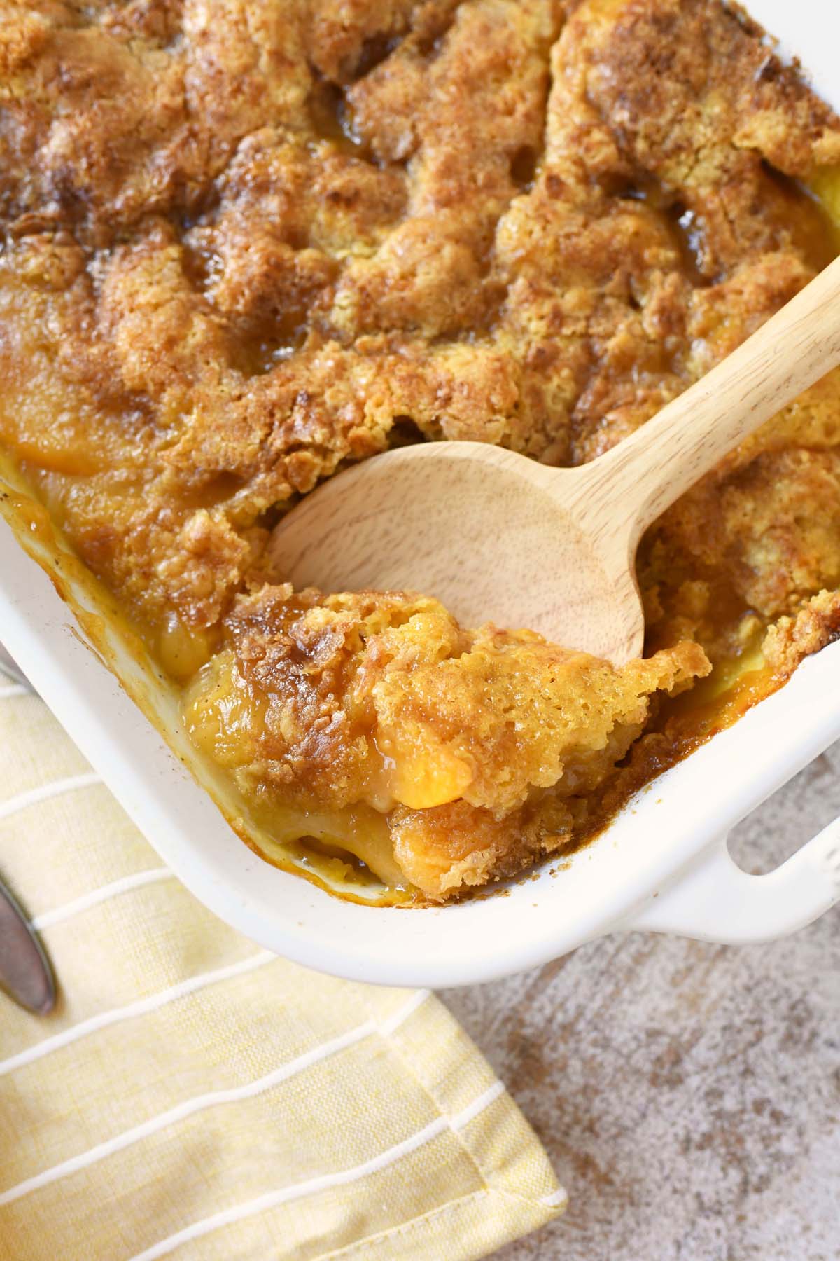 Peach Dump Cake in a white pan with a wooden spoon.