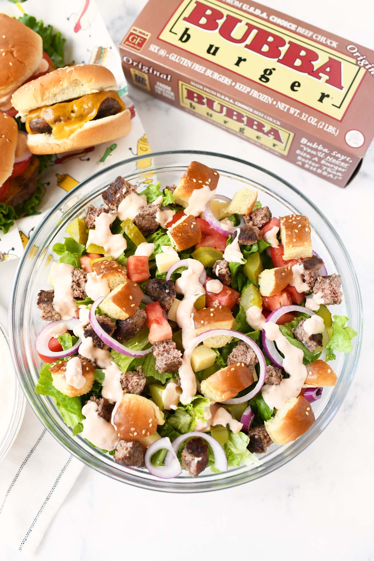 A large glass bowl of cheeseburger salad with a box of bubba burgers.
