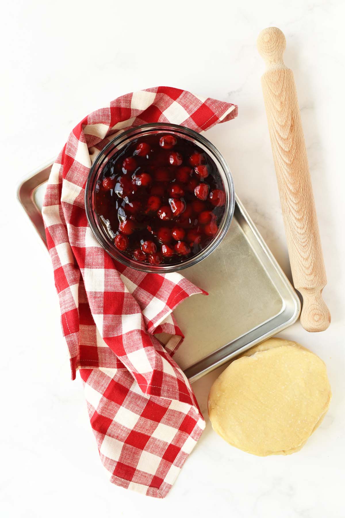 Cherry pie filling, rolling pin, dough, and checkered red napkin on a white table.