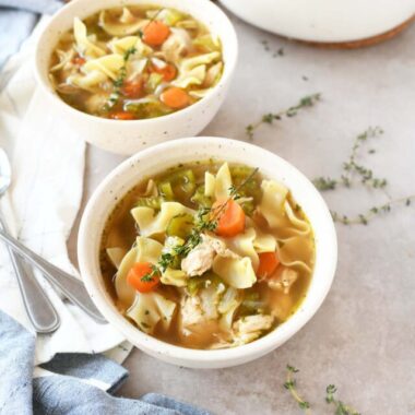 White bowls of chicken noodle soup with fresh thyme sprigs.