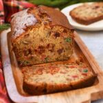 A sliced loaf of paradise banana fruitcake near a lighted Christmas tree, and flannel napkin.