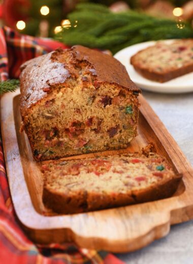 A sliced loaf of paradise banana fruitcake near a lighted Christmas tree, and flannel napkin.