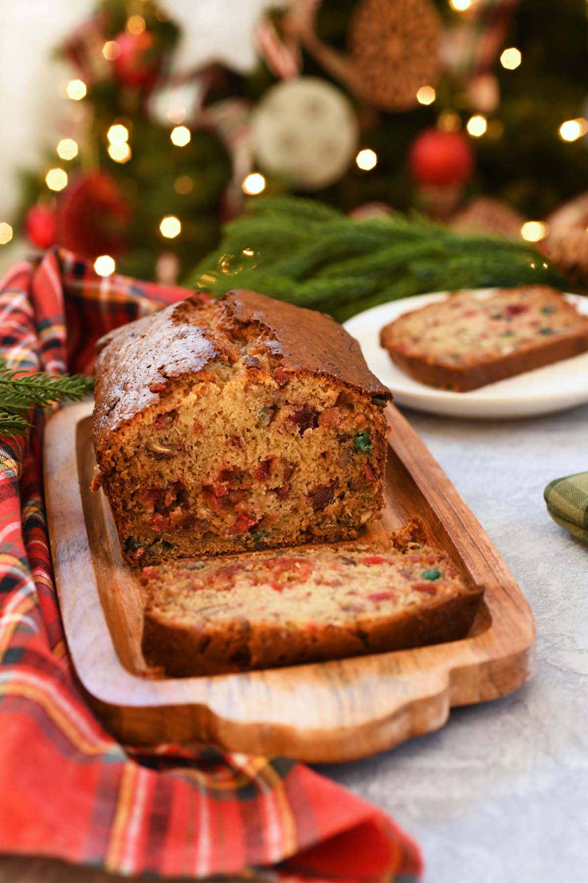 A sliced fruitcake loaf on a table with a lighted Christmas tree in the background.