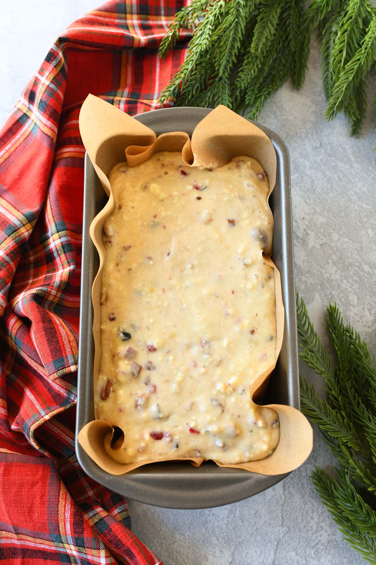 Banana bread fruitcake batter in a metal pan near a red, plaid napkin.