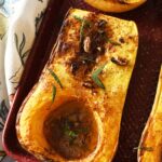 Bakd butternut squash halves with sage on a red baking sheet, near a floral napkin.
