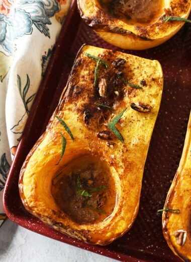 Bakd butternut squash halves with sage on a red baking sheet, near a floral napkin.