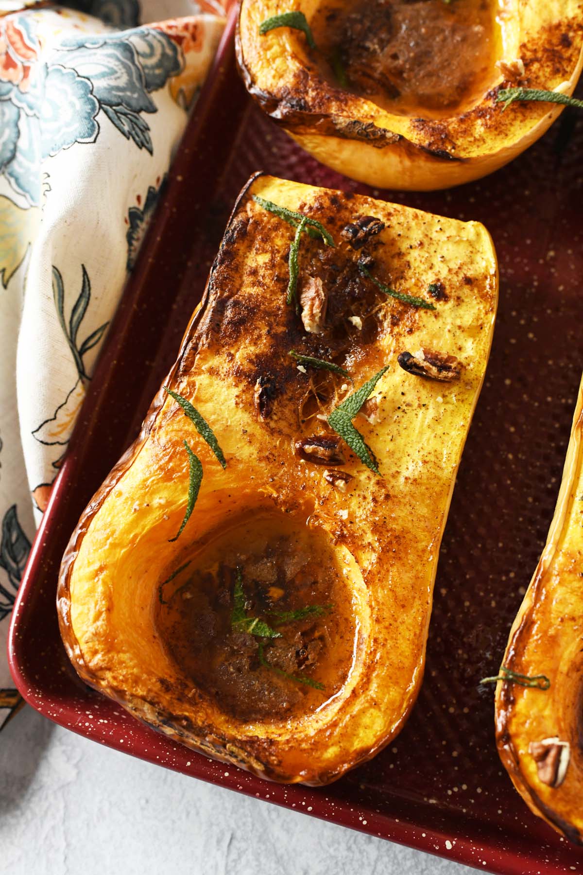 Baked butternut squash halves with sage on a red baking sheet, near a floral napkin.