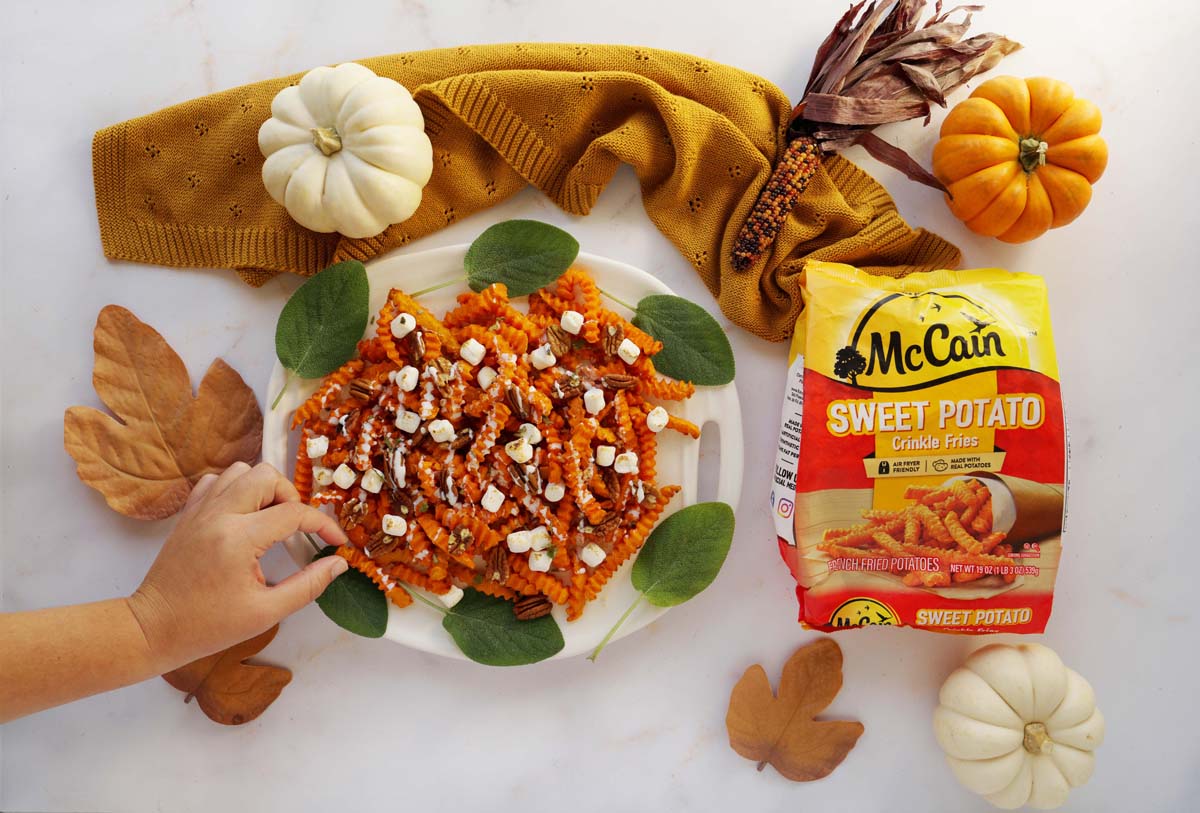 A hand grabbing a loaded McCain Sweet Potato fry with marshmallow and pecans on it. Mini pumpkins are nearby on the marble surface.