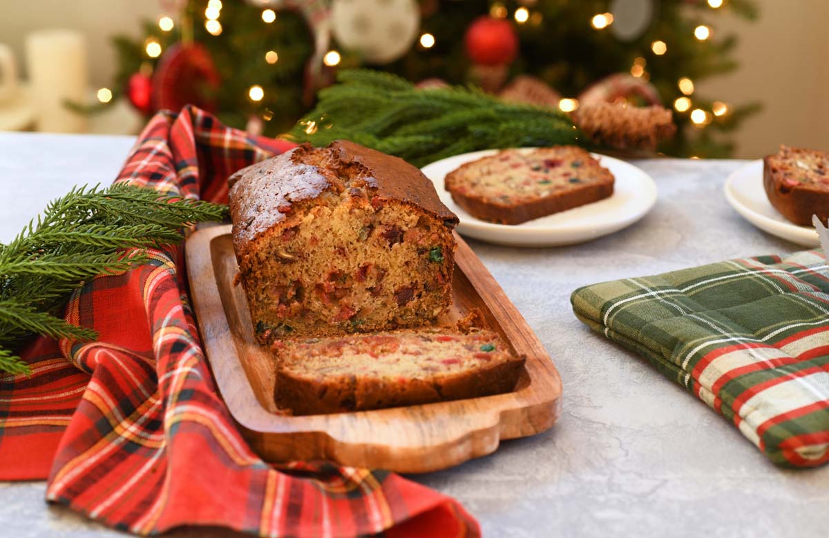 A sliced fruitcake with slices on little plates on a Christmasy setting with a lighted tree in the background.