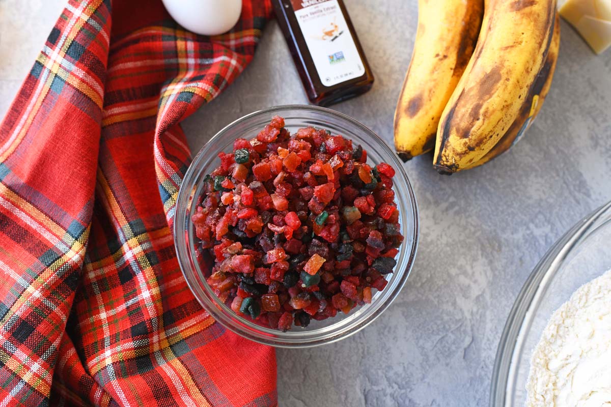 A bowl of crystalized Paradise Fruit Mix near banana bread ingredients and a red, flannel napkin.