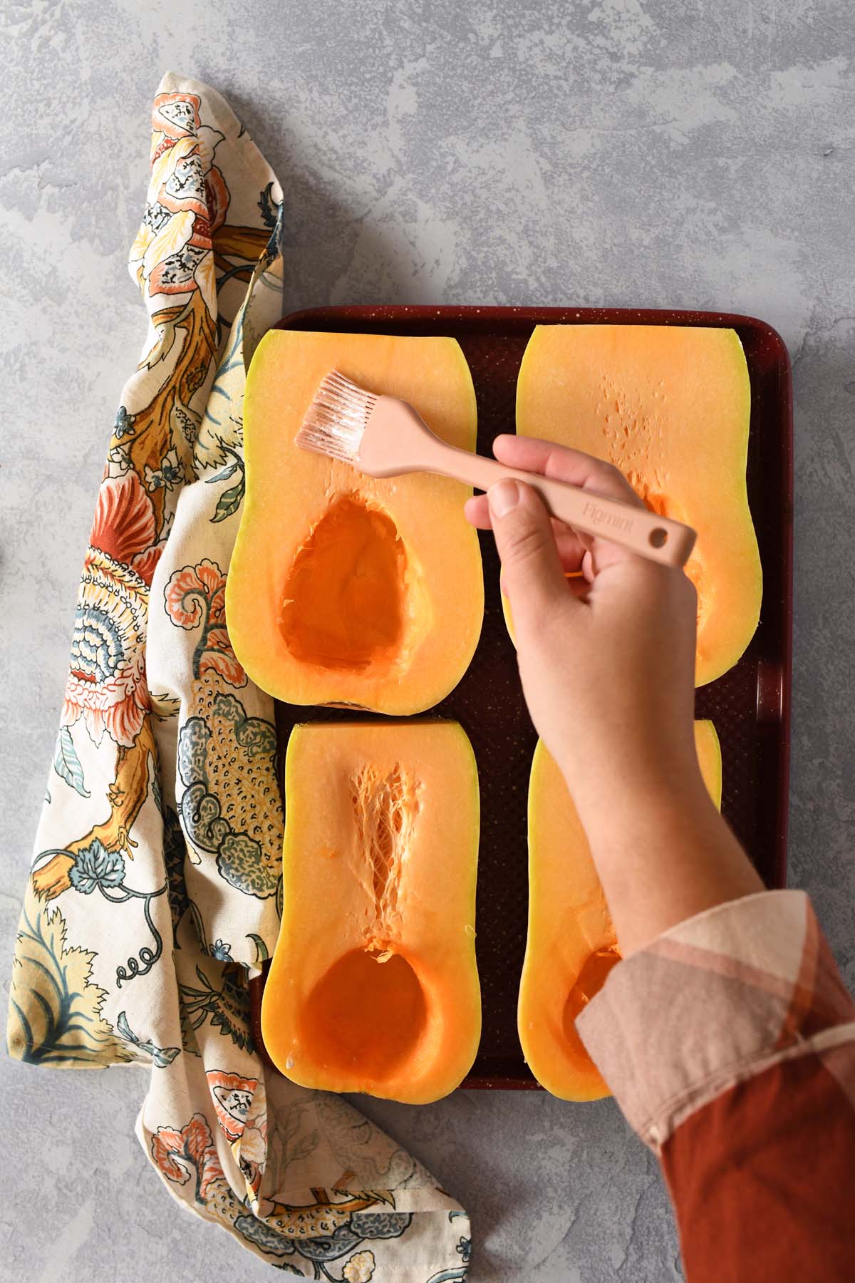 A hand brushing butter on butternut squash halves. They are on a red baking sheet near a floral napkin.