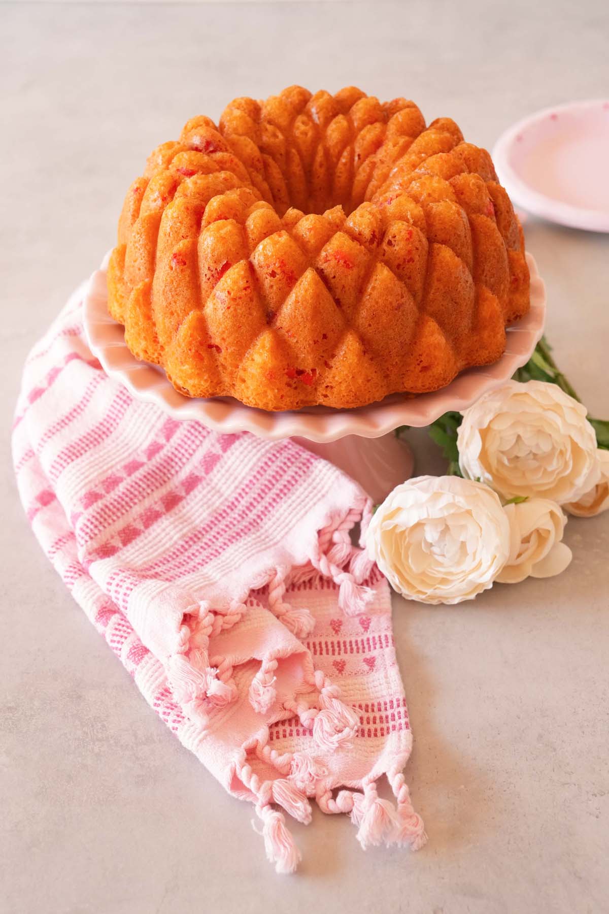 Cherry kaleidoscope bundt cake on a pink cak stand near a pink napkin and white flowers.