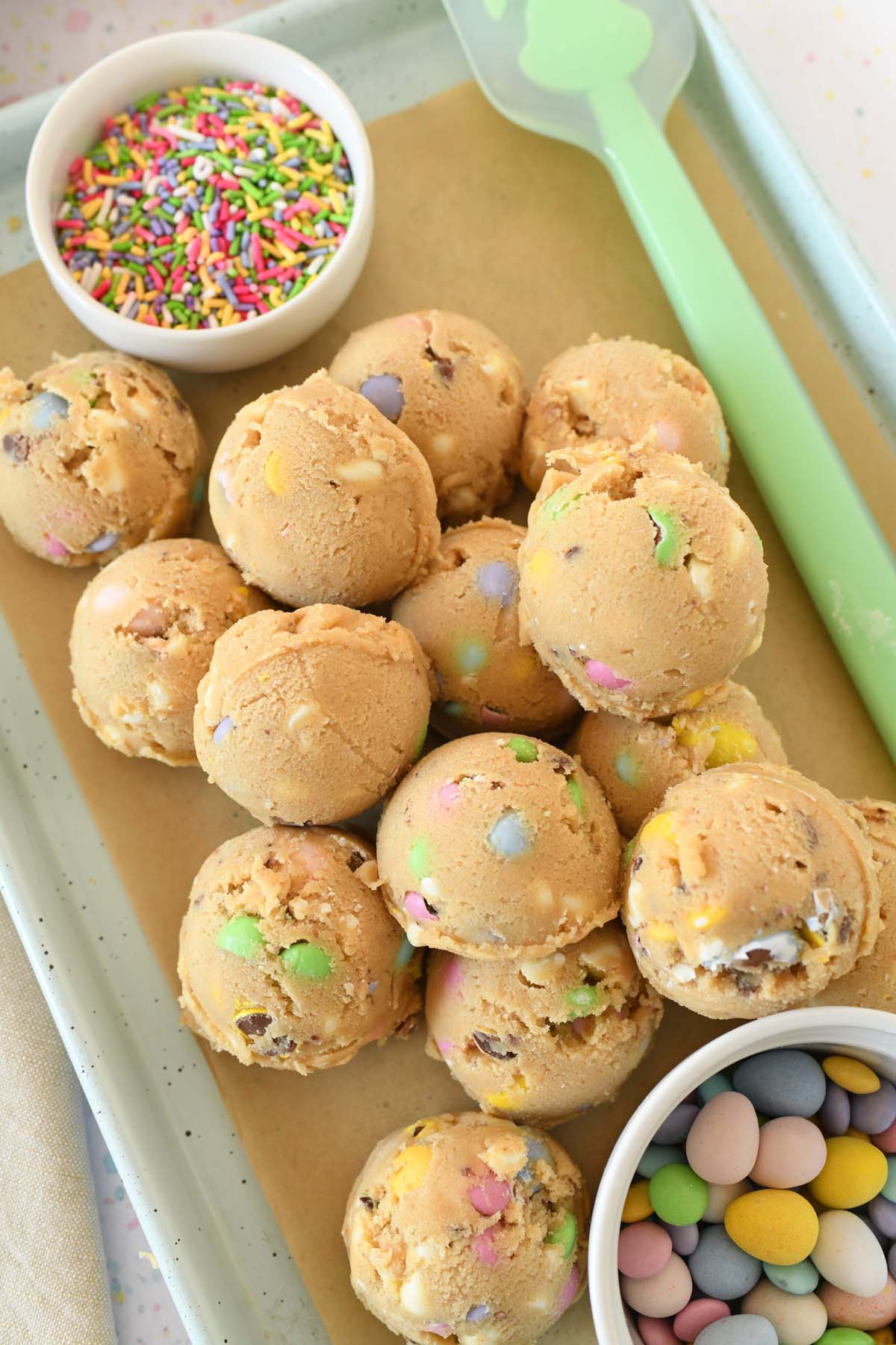 Up-close shot of unbaked cookie dough balls on a parchment-lined baking sheet and pastel candies, and sprinkles.