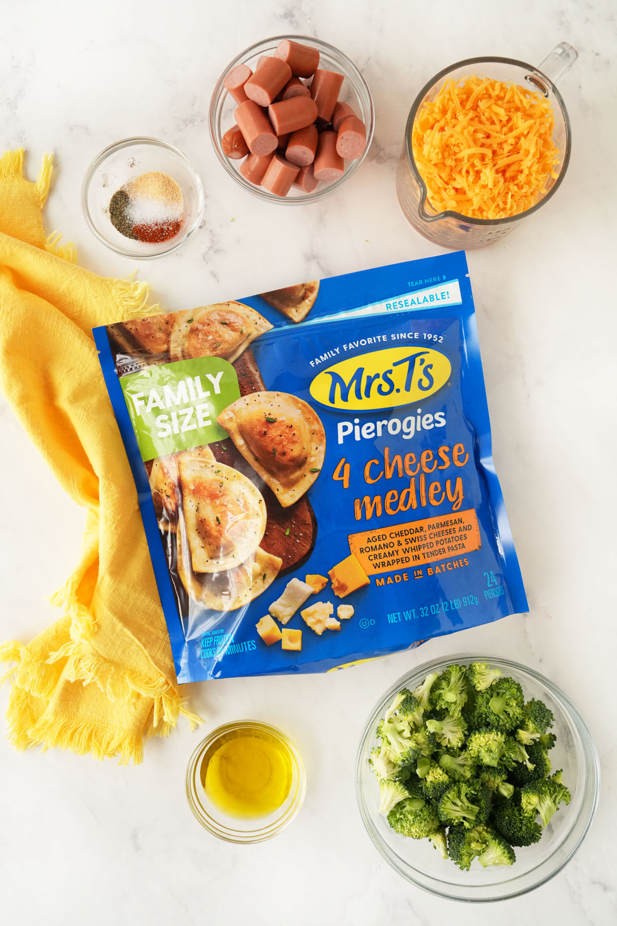 Mrs Ts family size four cheese medley, spices, hot dogs, cheese, broccoli, oil, and a napkin on a white surface.