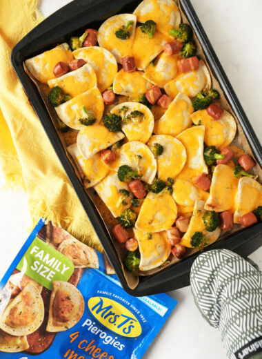 An oven-mitted hand holding a cheesy sheet pan of pierogis and hot dogs near a bag of pierogis and a yellow napkin.