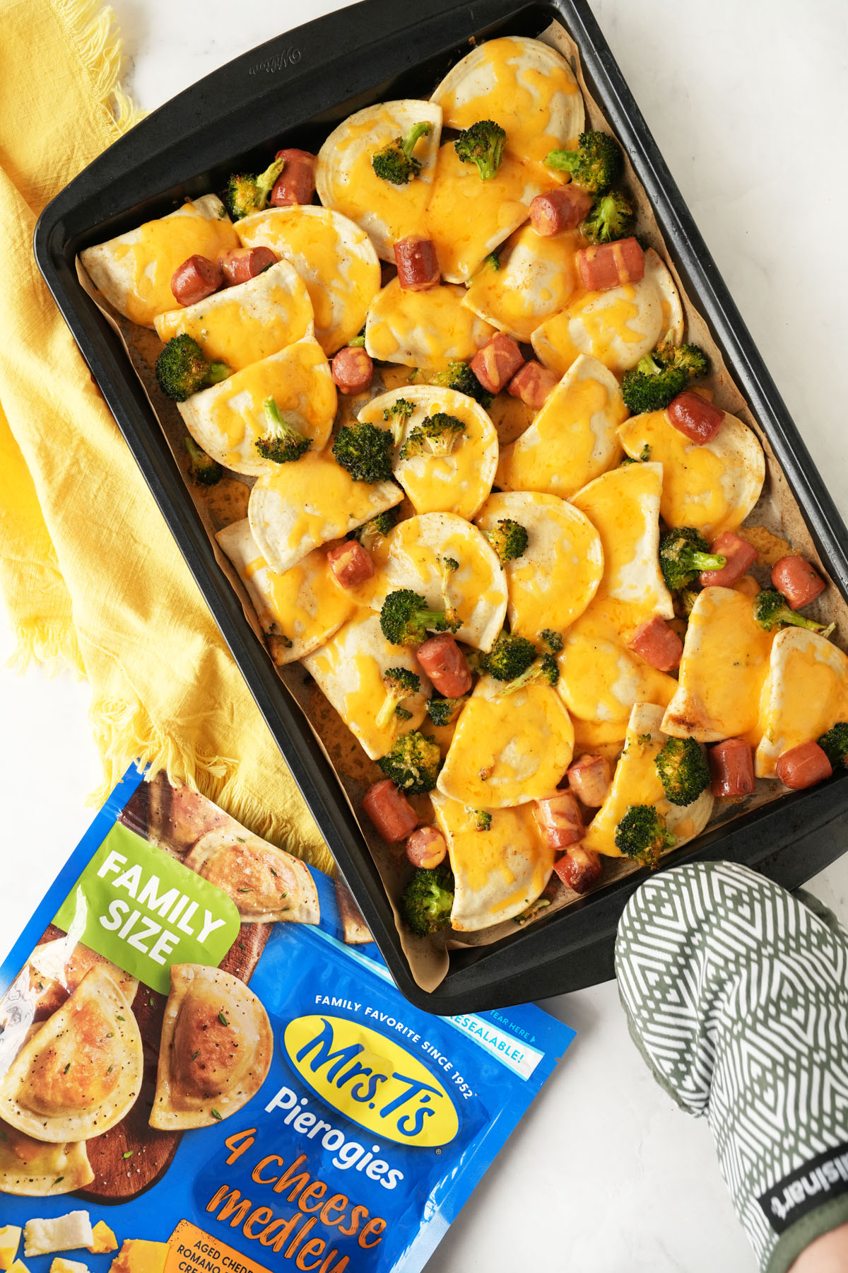 An oven-mitted hand holding a cheesy sheet pan of pierogis and hot dogs near a bag of pierogis and a yellow napkin.