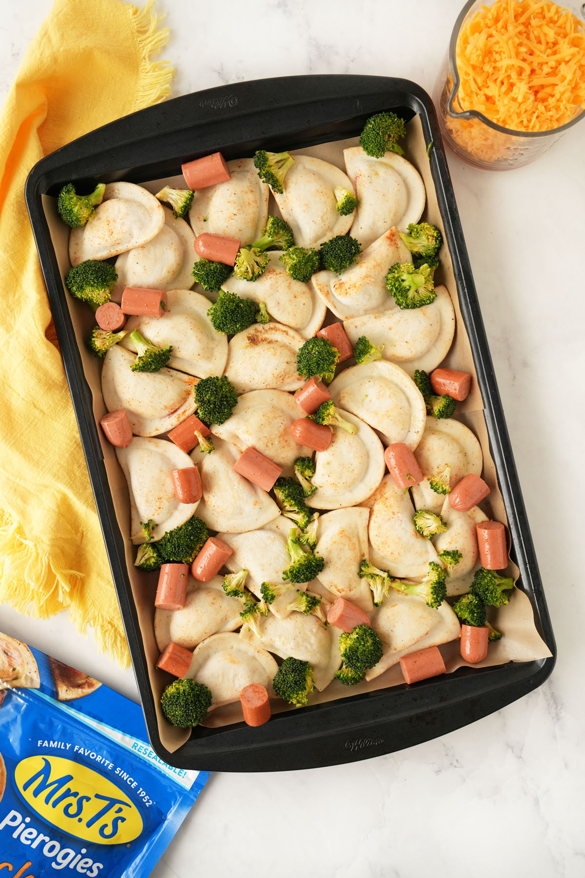 Seasoned pierogis, broccoli, and hot dogs on a sheetpan near cheese, a yellow napkin, and product bag.