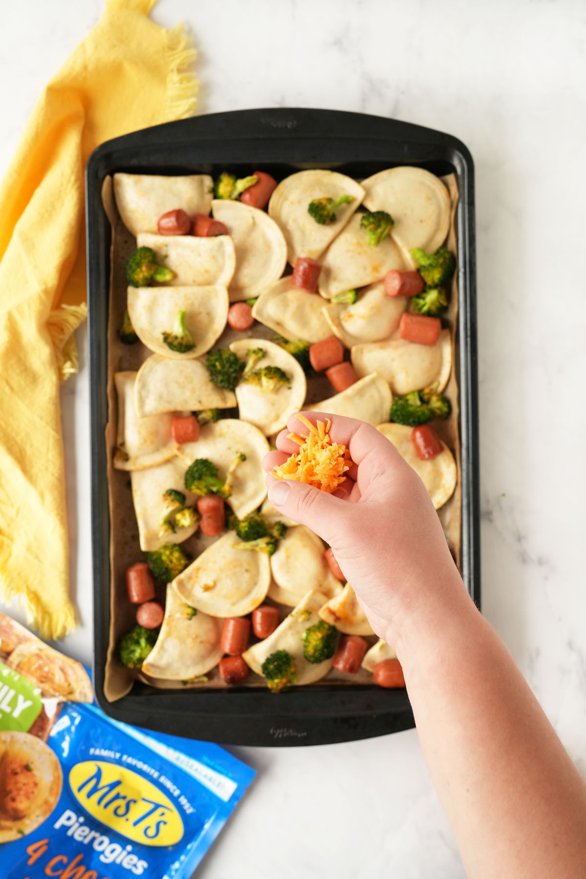A hand ready to sprinkle cheese on a pan of baked pierogis with hot dogs and broccoli.
