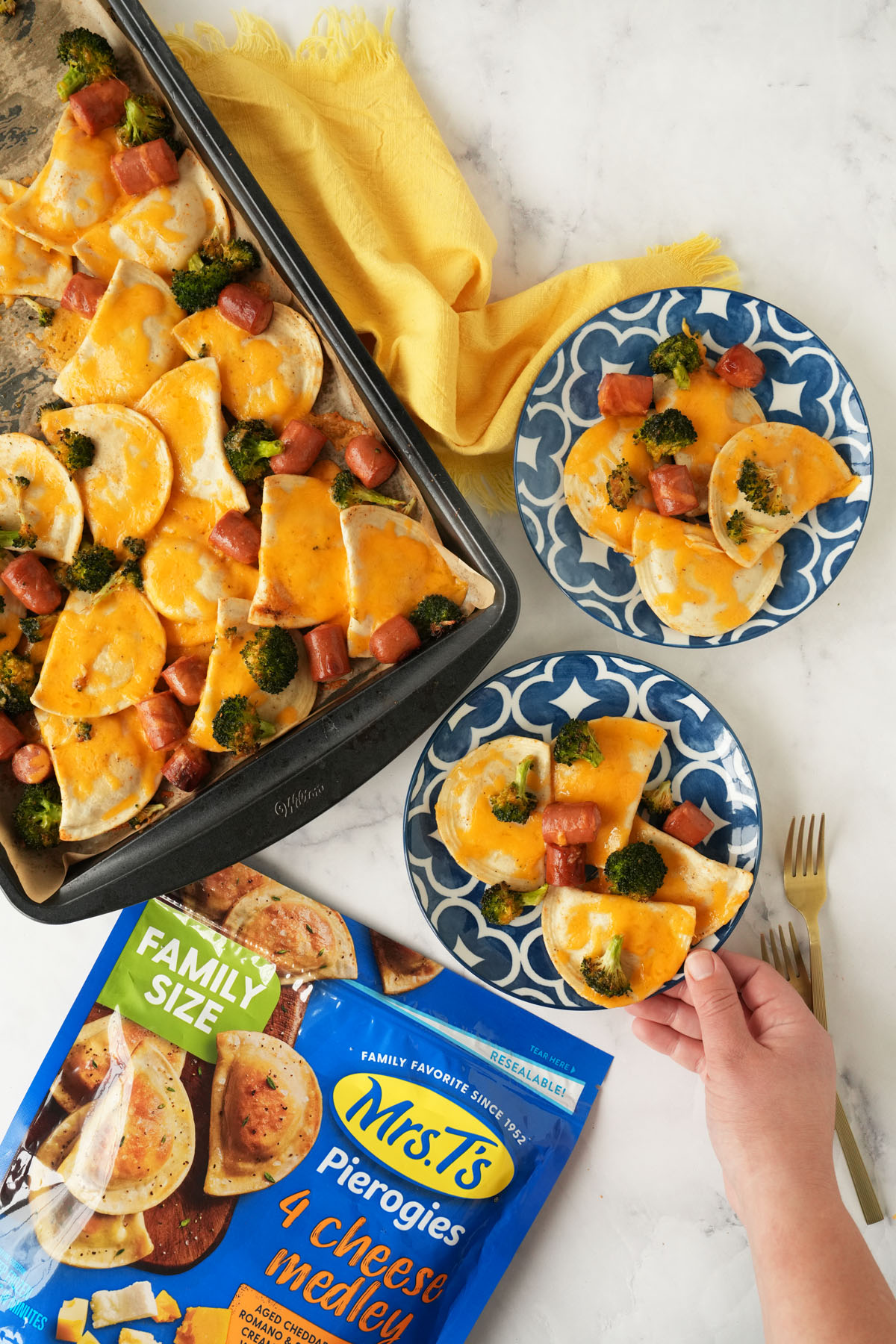 Two plates of sheet pan pierogis near a sheetpan of pierogis, a yellow napkin, pierogis packge, and hand.