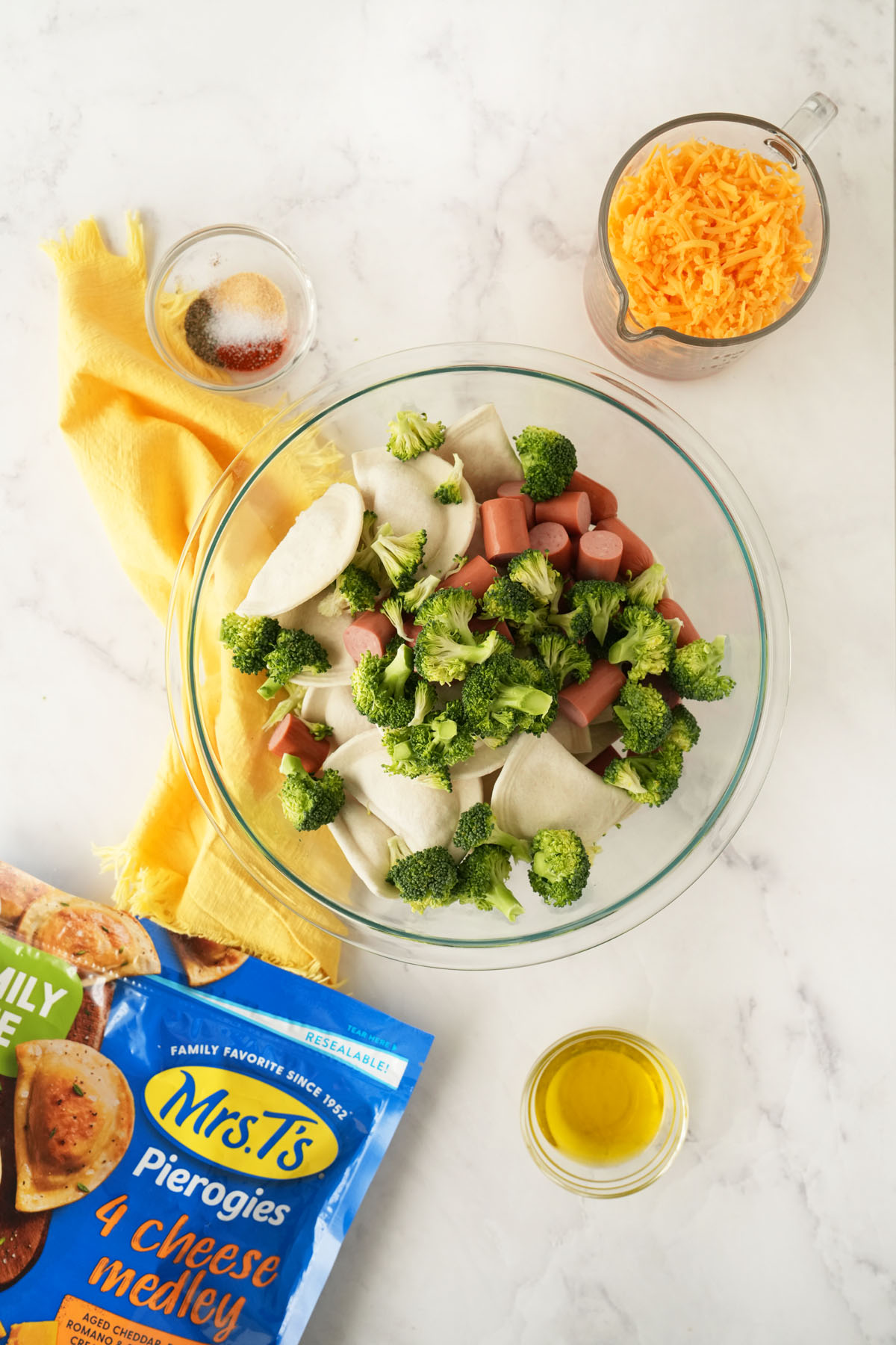 Broccoli, hot dogs, and pierogis in a glass bowl with seasonings, cheese, oil, and an extra pierogis bag nearby.