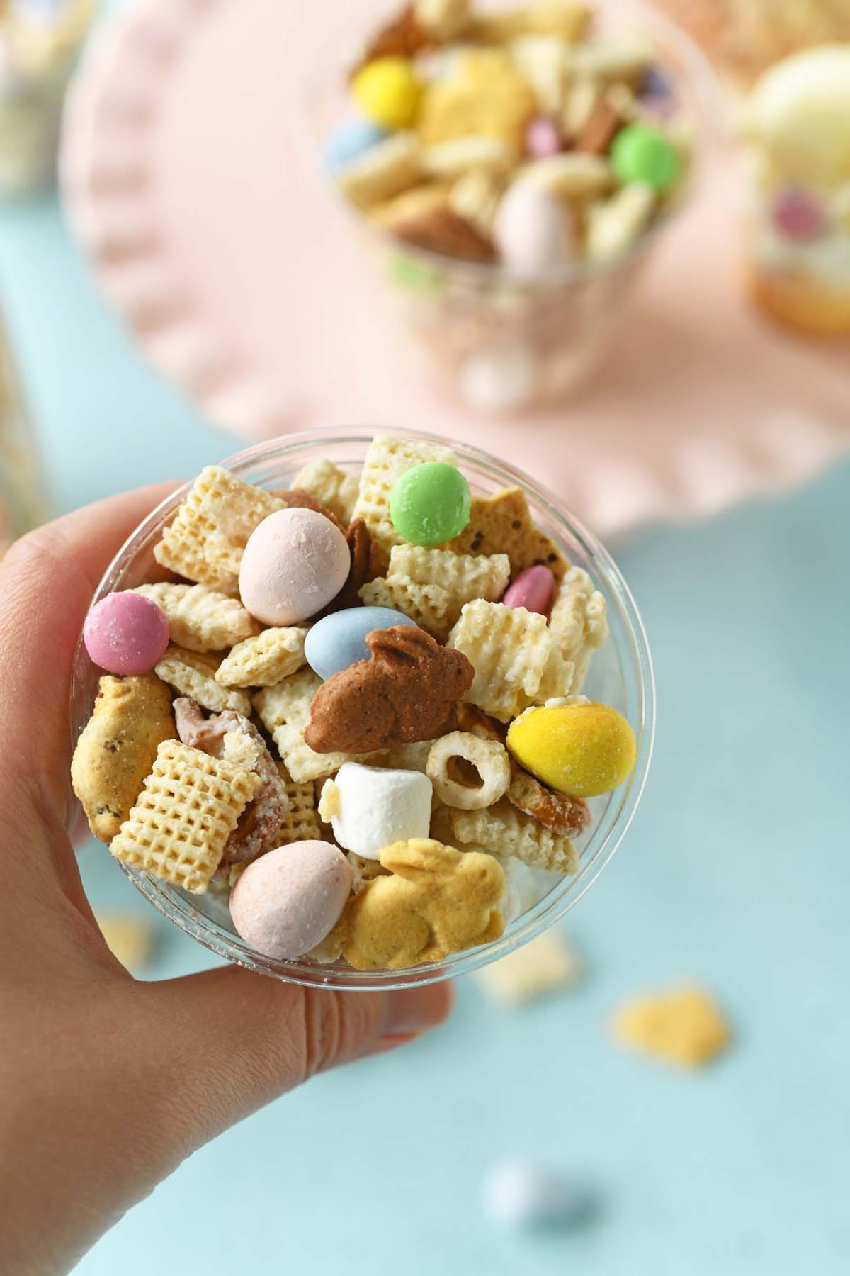 A hand holding up a cup of colorful bunny bait snack mix with sweet and savory elements.