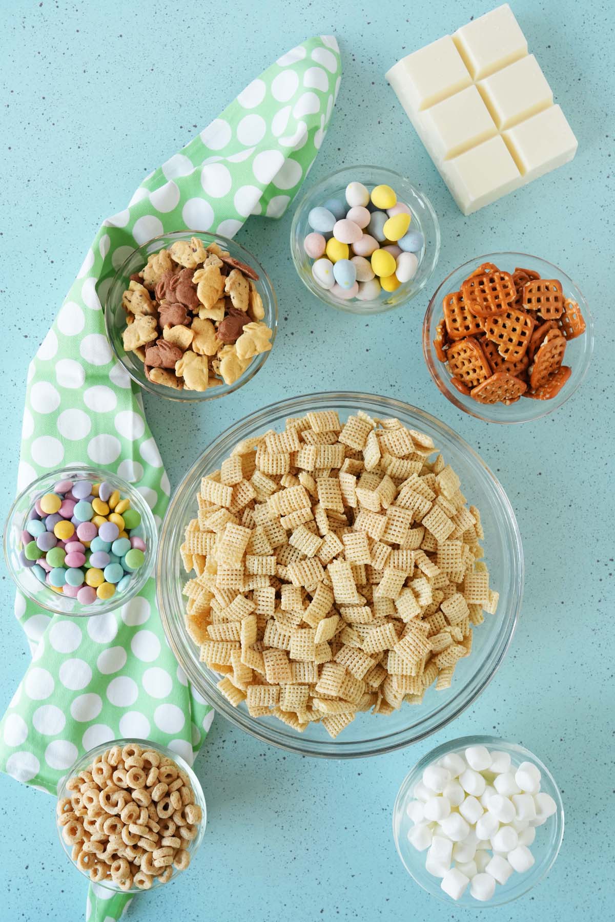 Ingredients for bunny bait separated into assorted sized glass bowls, including mini marshmallows, candies, pretzels, and cereals.