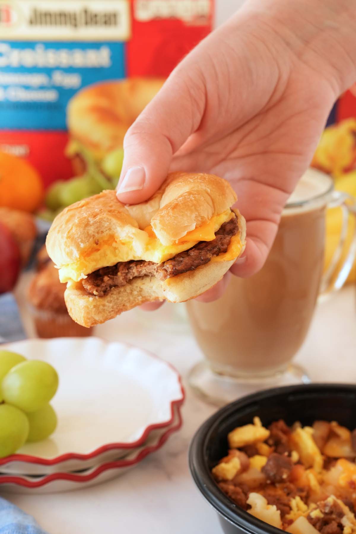 A bitten Jimmy Dean Breakfast Crossaint in a hand near other breakfast foods.