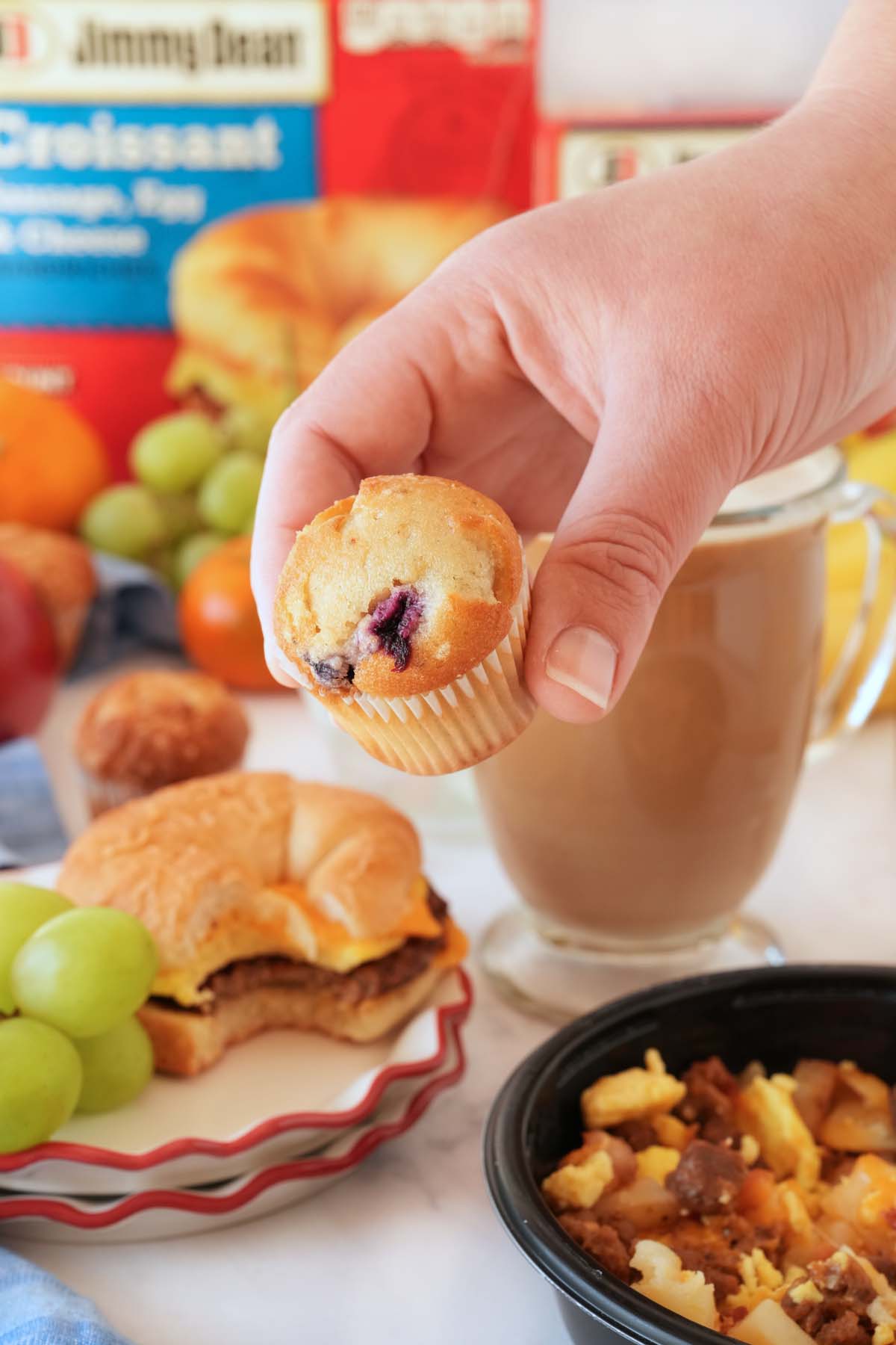 A hand holding a mini blueberry muffin up close in a breakfast setting.