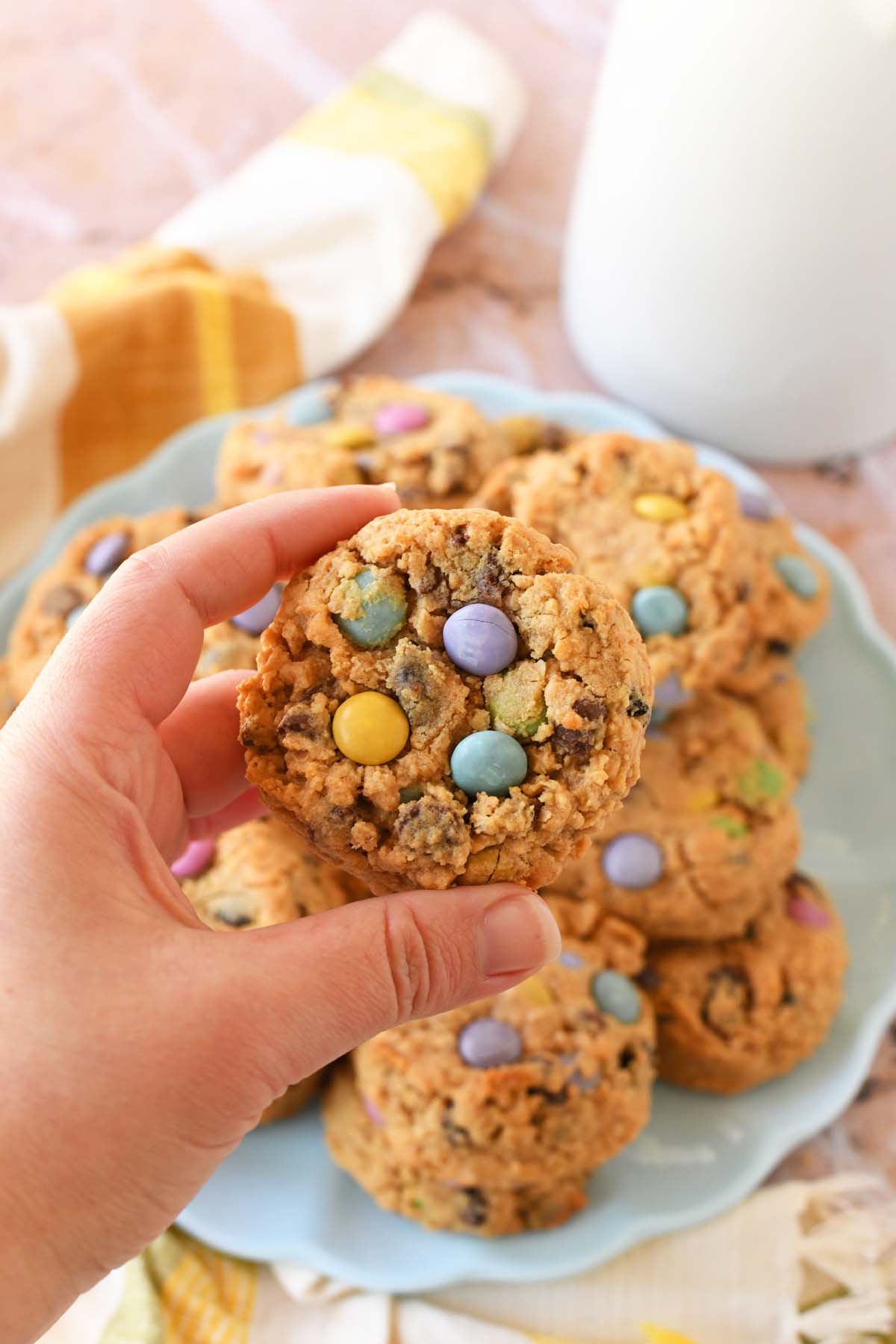 A hand holding a pastel monster cookie above a plate of monster cookies.