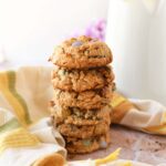 A stack of golden-brown spring monster cookies near a striped napkin,and pitcher with a flower inside.