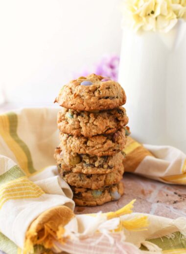 A stack of golden-brown spring monster cookies near a striped napkin,and pitcher with a flower inside.