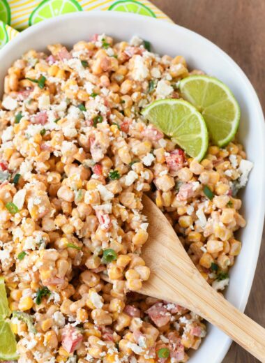 Colorful smoky elote salad with green lime wedges and a wooden spoon.