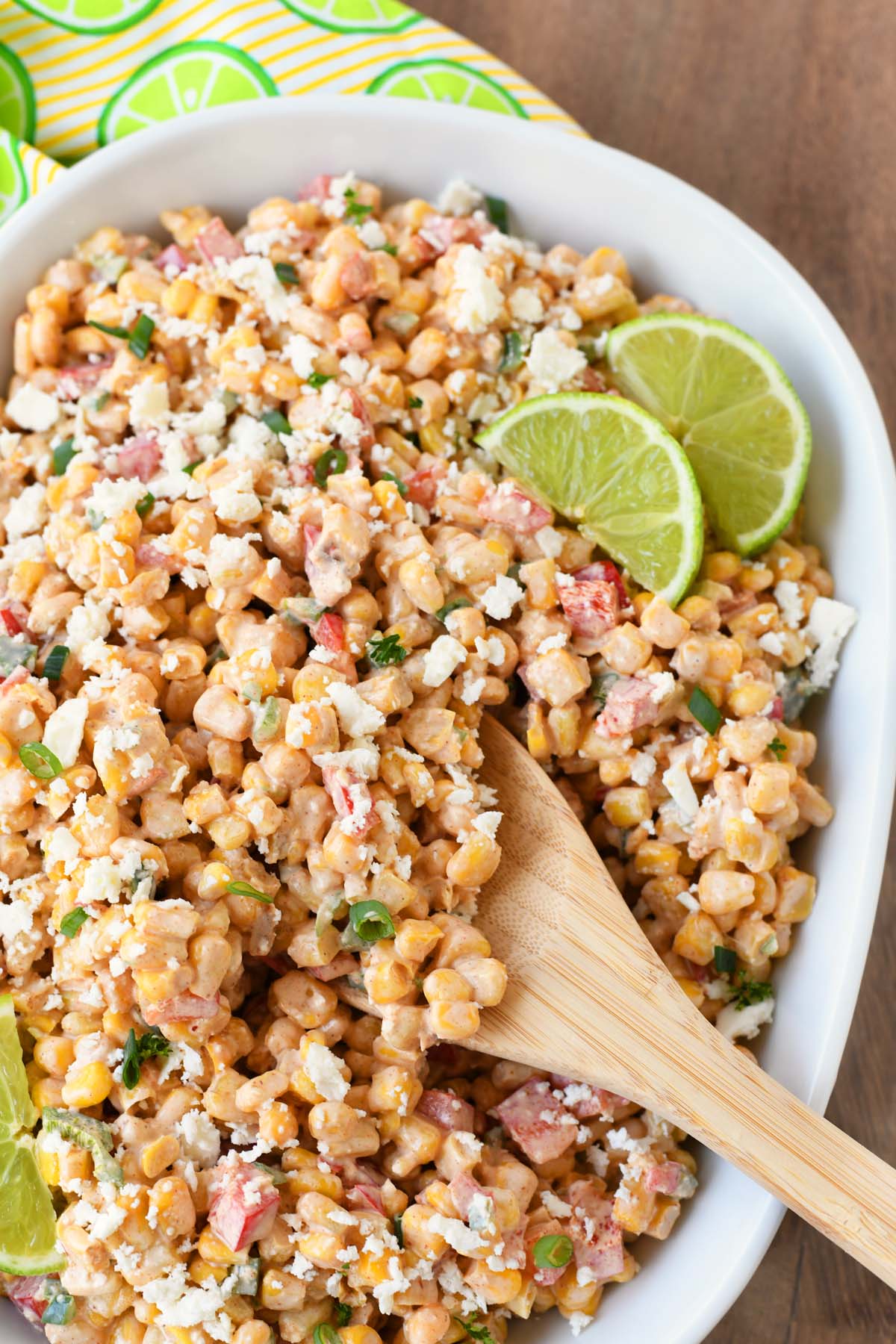 A wooden spoon resting inside of corn salad near sliced limes.