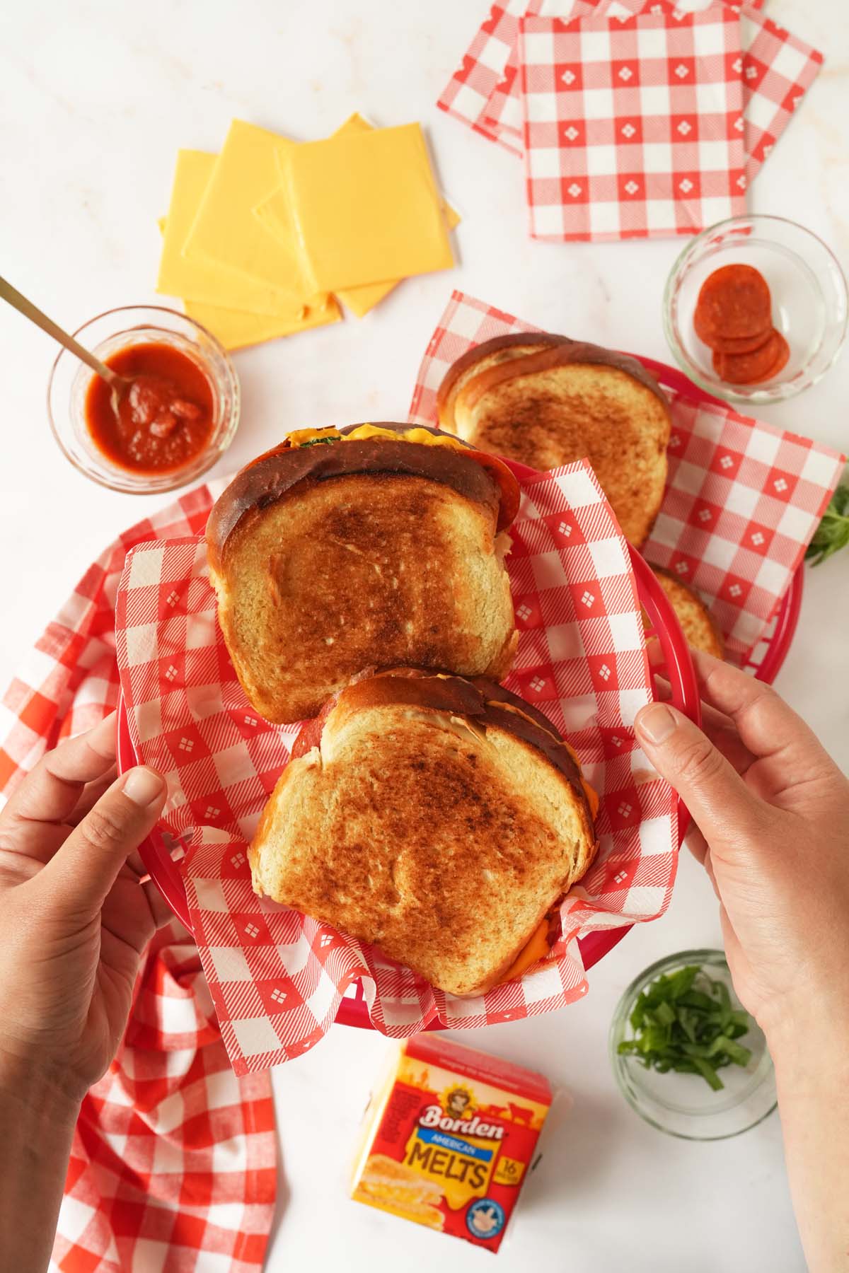 Tan hands holding up a red, checkered basket with golden-brown grilled cheese sandwiches inside.