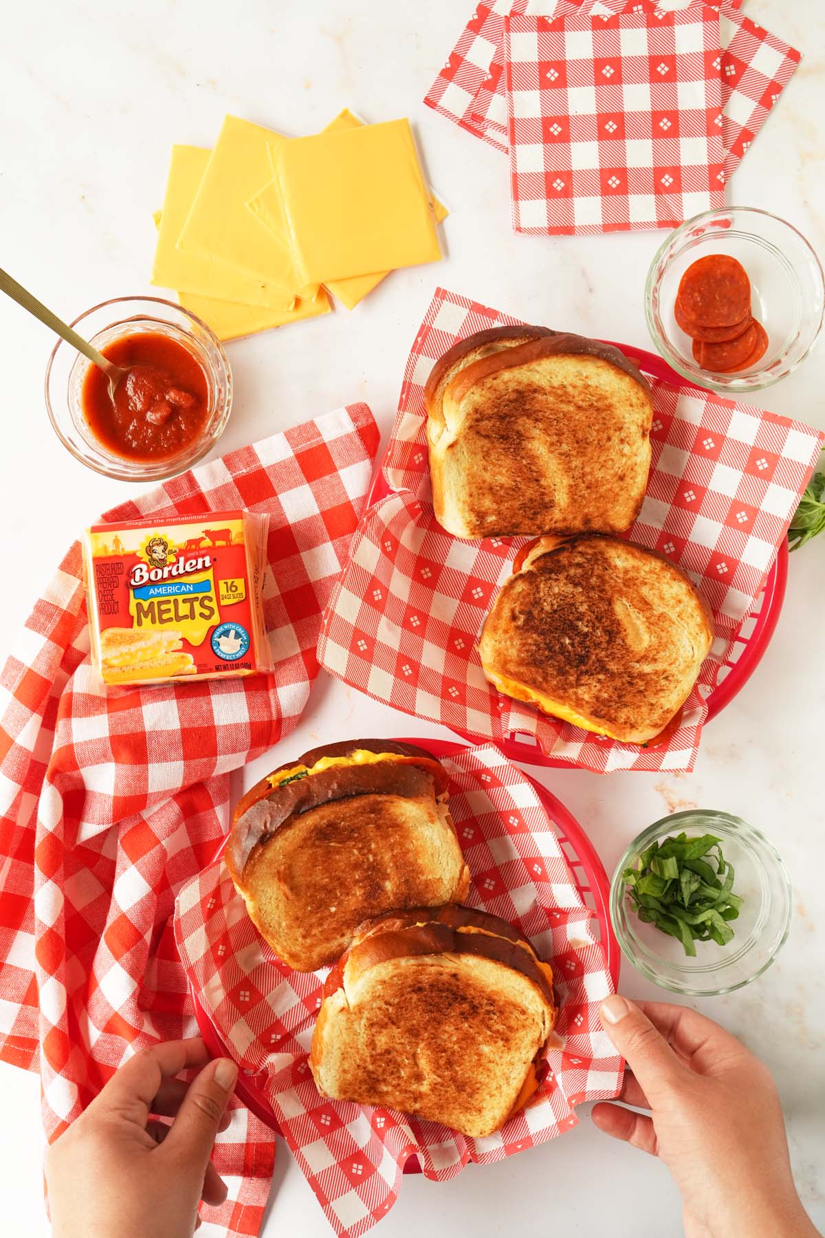 A hand touching a basket of golden-brown grilled cheese sandwiches with more sandwiches and ingredients nearby.