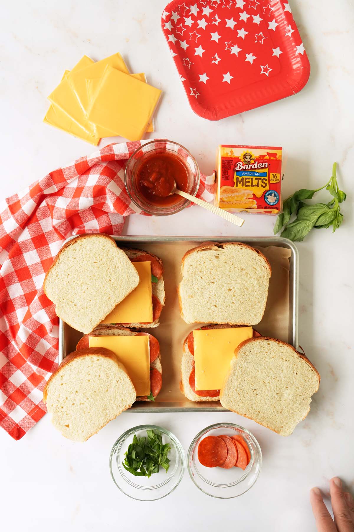 Uncooked pepperoni pizza grilled cheese sandwiches on a white table with a red checkered napkin and ingredients nearby.