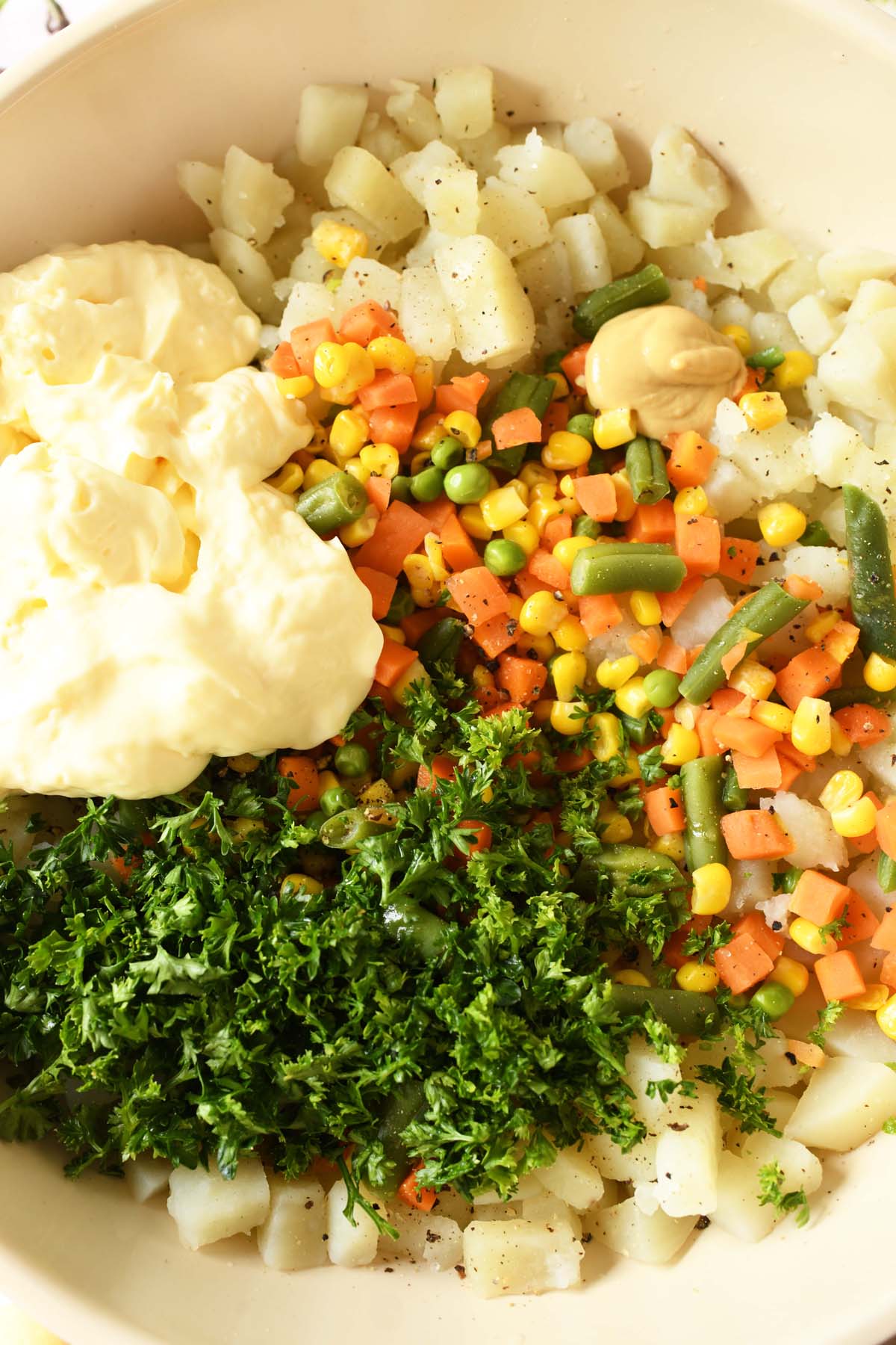 Brazilian potato salad ingredients like mixed veggies, mayo, spices, parsley, and potatoes in bowl up-close.