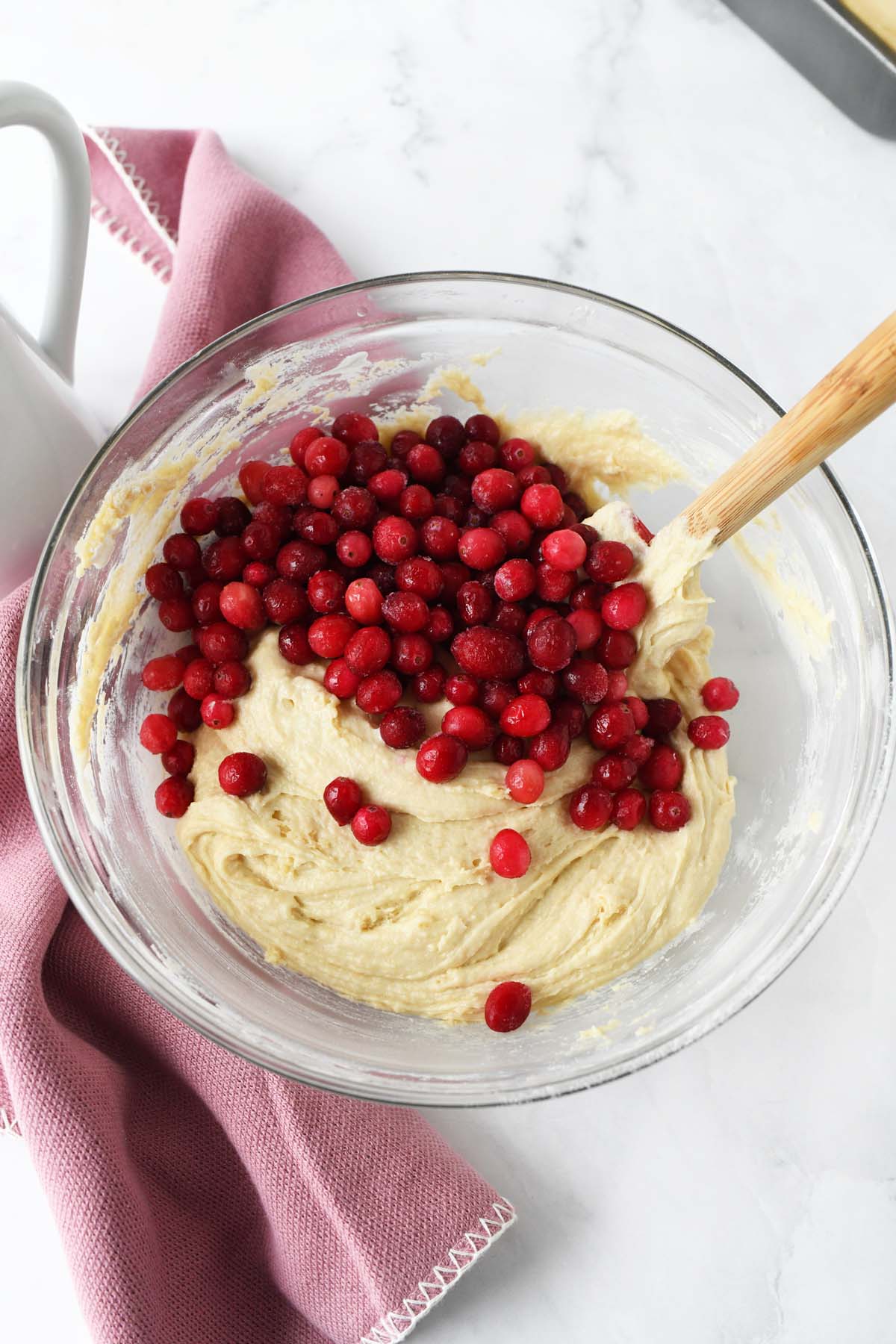 Frozen cranberries in batter in a glass mixing bowl near a pink napkin.