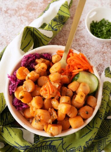 A gold fork resting inside a bowl of pickled, colorful veggies and rice topped with orange gochujang scallops.