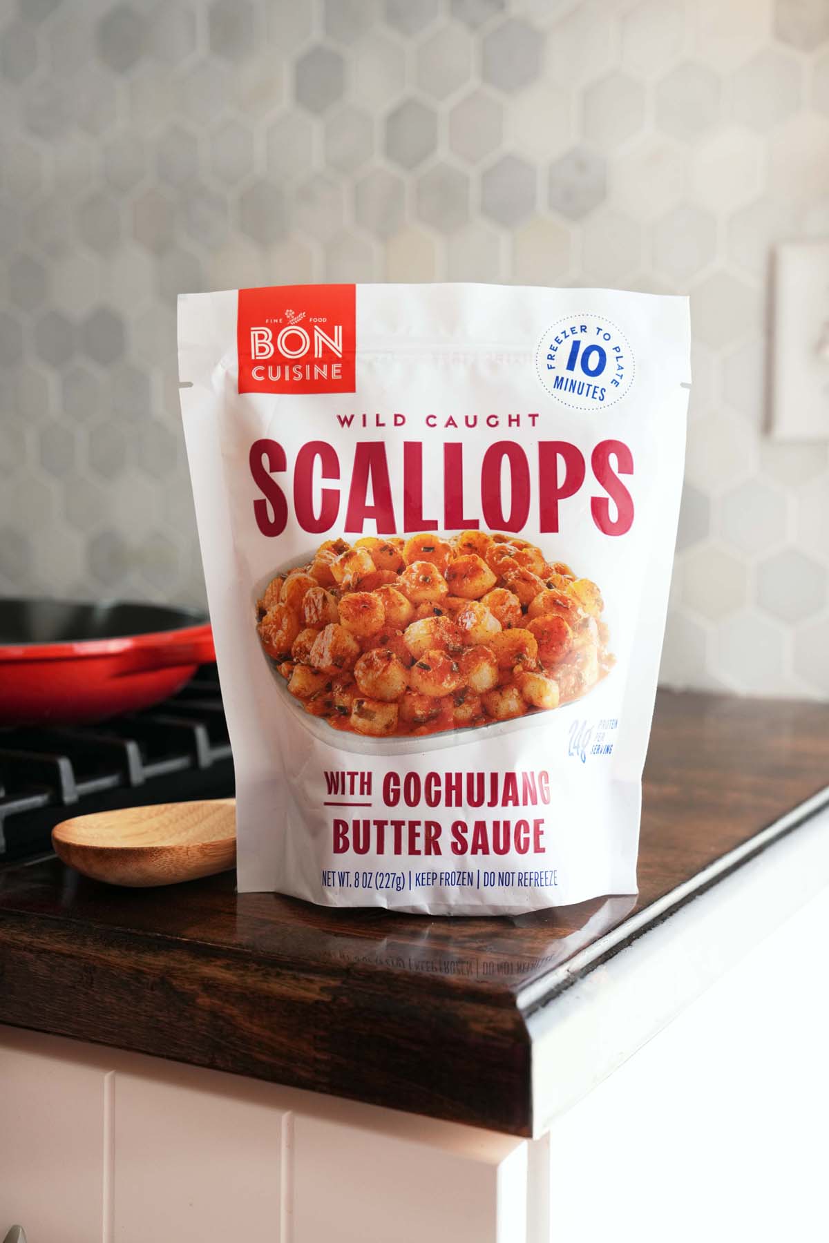 A bag of Gochujang scallops on a wooden counter near a red skillet, and wooden spoon.