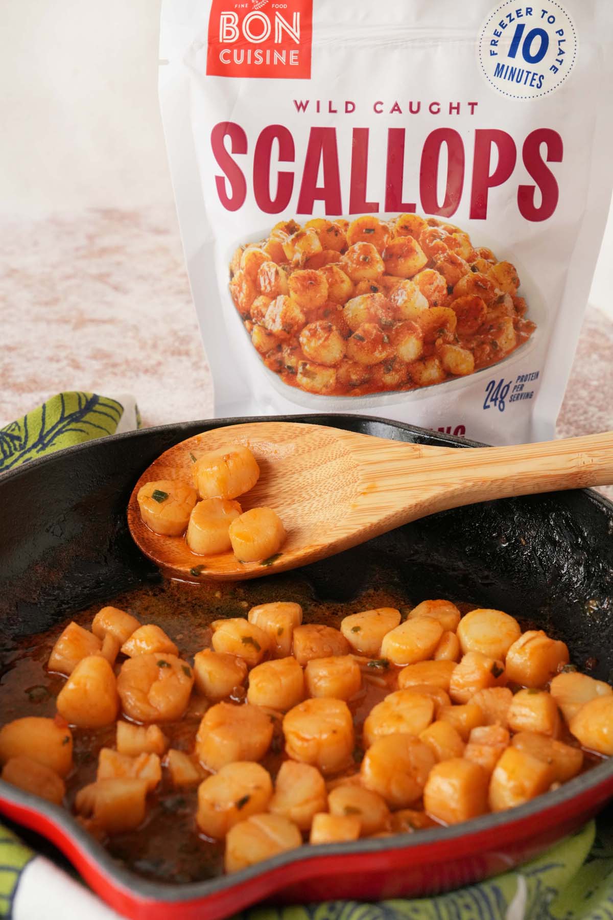 Orange gochujang scallops in a black and red skillet near a wooden spoon, and bag of scallops.