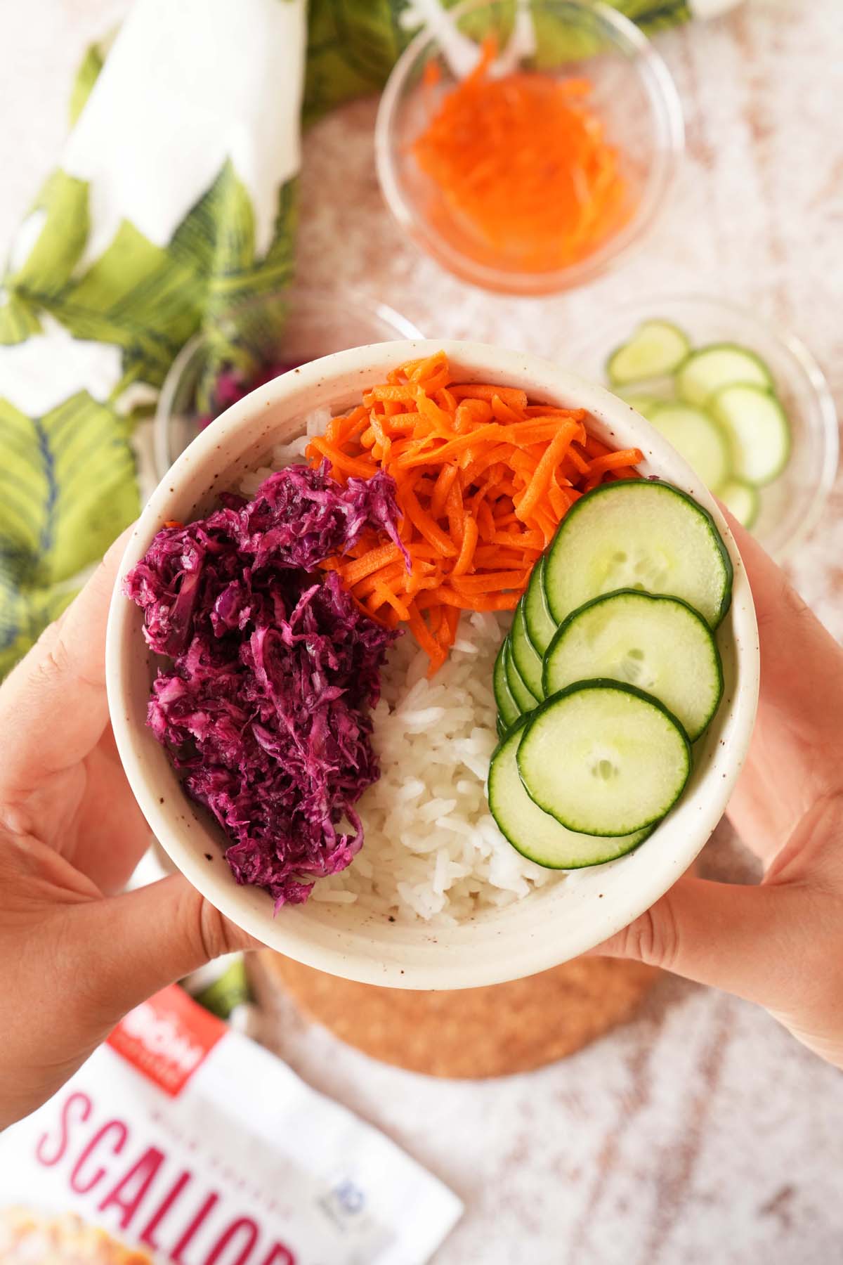 Hands holding up a colorful rice bowl of pickled red cabbage, cucumbers, and carrots.