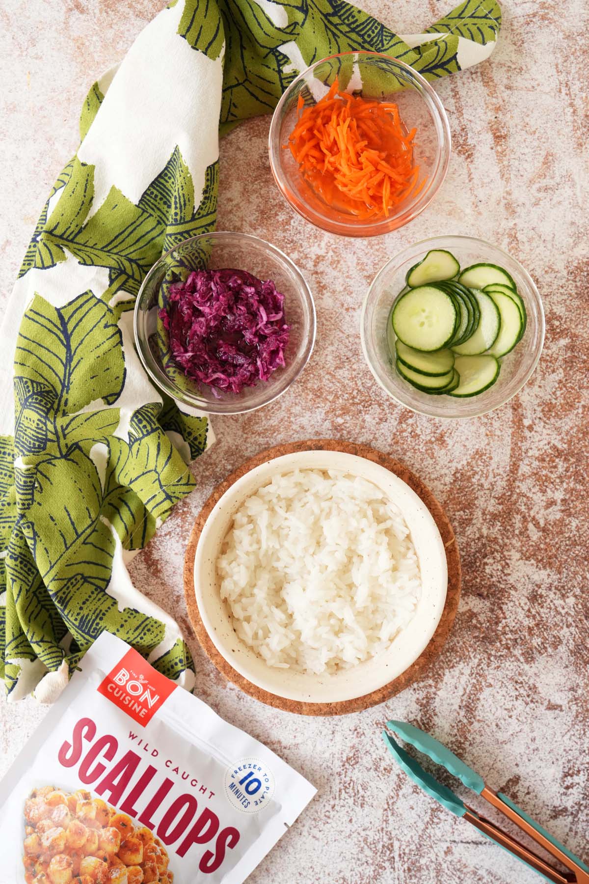 Colorful pickled veggies for rice bowls on a tan, textured surface with a green napkin, and a bag of scallops nearby.