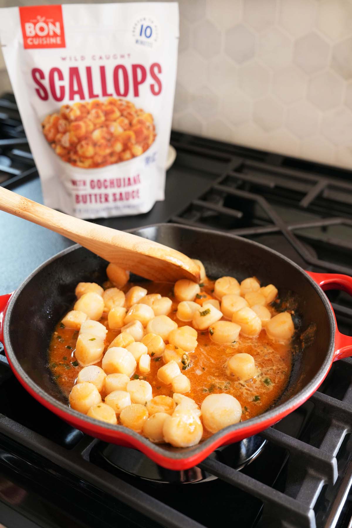 A red skillet with Gochujang scallops inside near a wooden spoon. A bag of Gochujang Bon Cuisine Scallops is in the background.